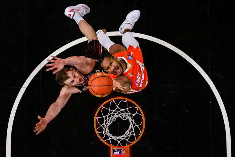 Aerial view of two basketball players, one in black and one in orange, jumping to reach the ball above the hoop during a game, both extending their arms towards the ball.