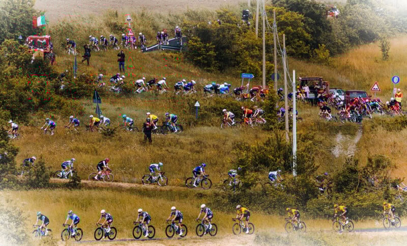 A large group of cyclists ride along a winding road through grassy fields, with support vehicles and spectators nearby. The scene is lively and colorful, with trees and flags in the background.