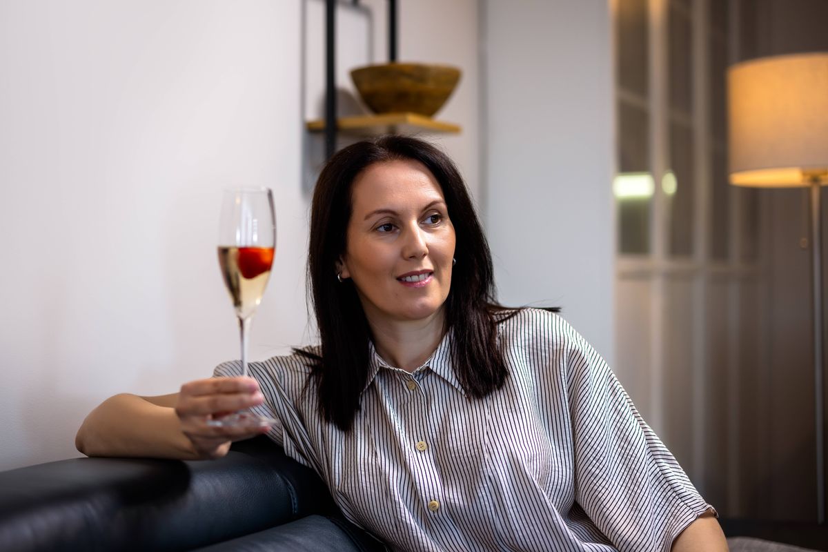 A sophisticated woman in her 40s relaxing at home with a glass of wine and a fresh strawberry. She is sitting comfortably, expressing happiness and indulgence in a peaceful domestic atmosphere.