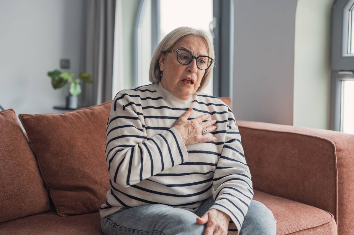 Senior adult woman sitting on a couch suffering from sudden chest pain and shortness of breath, indicating possible heart problems or anxiety attack during a health crisis at home