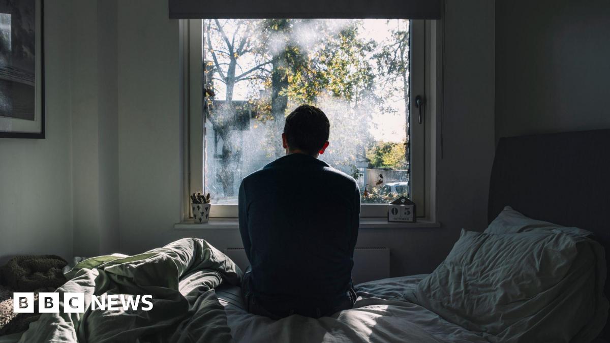 A man looks out of a window. He is in a dark room on an unmade bed. The atmosphere seems negative.