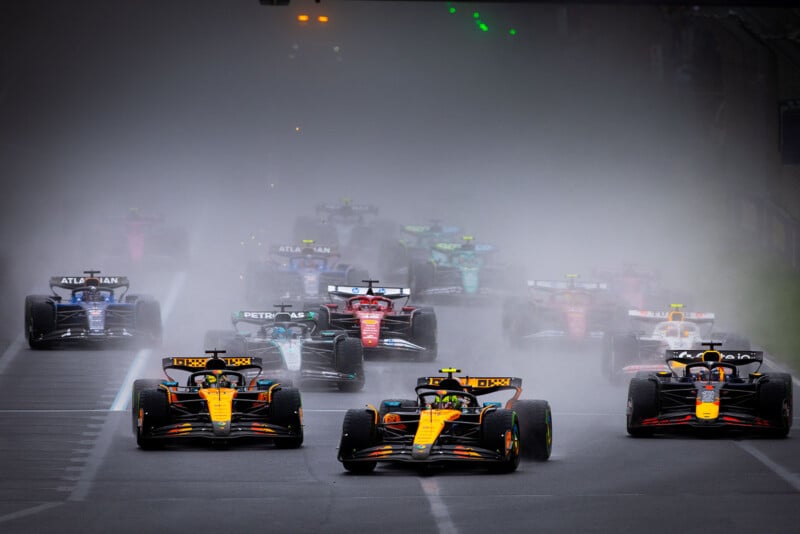 Formula 1 cars race through heavy spray on a wet track, with two orange and black cars leading the pack, followed by cars in various colors barely visible in the mist behind them.
