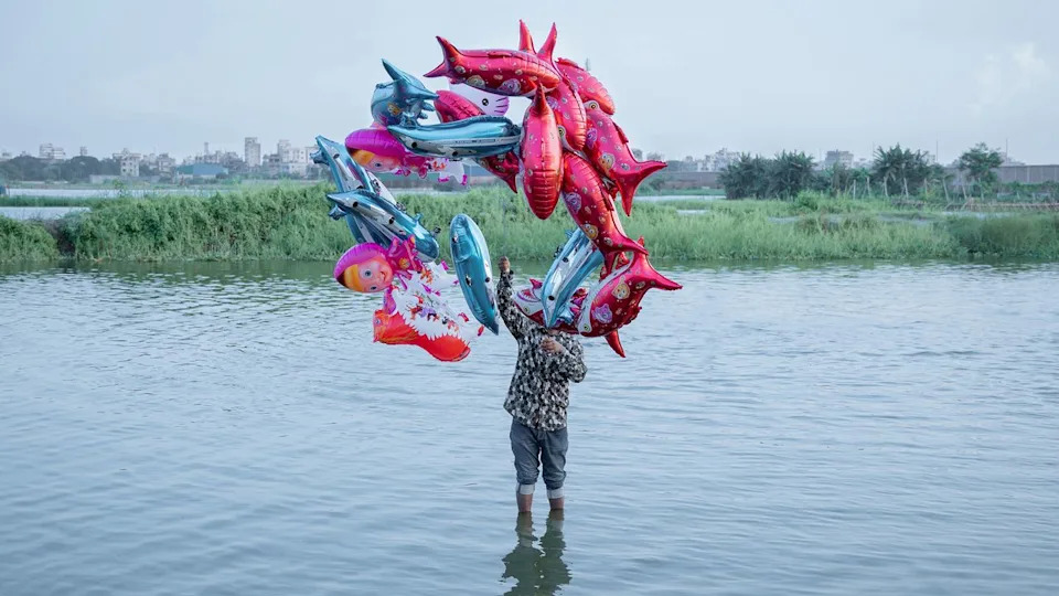 The fish belong in the river, but the river is gone. A man stands in the water, while balloons shaped like dolphins float above, a parody of joy.