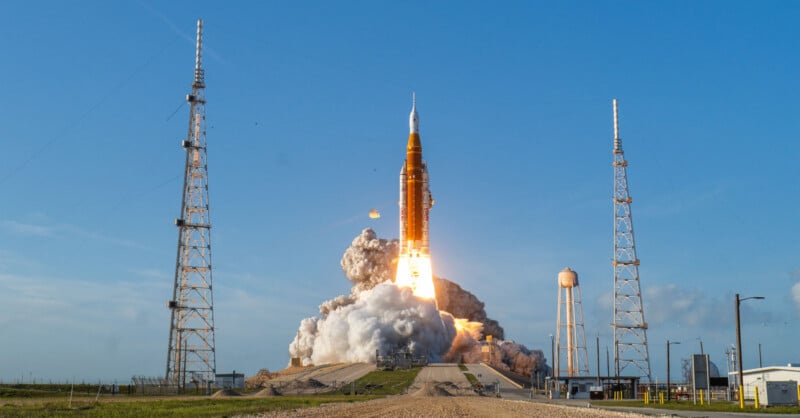A large rocket launches from a pad, emitting bright flames and smoke, with blue sky overhead and metal towers and a water tower surrounding the launch site.