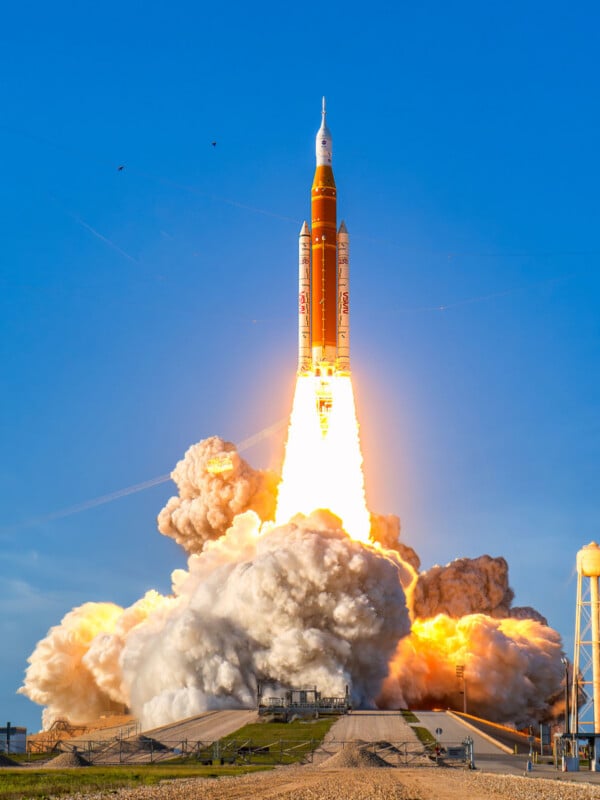 A rocket launches into a clear blue sky, releasing bright flames and thick clouds of smoke from its engines as it lifts off from a launch pad.