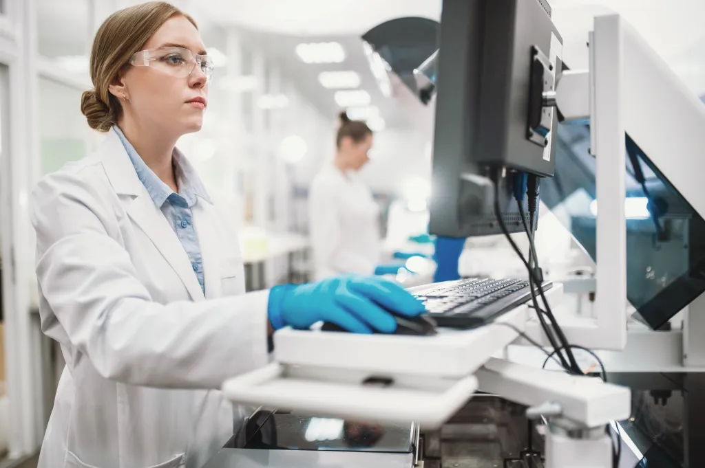A woman in a lab coat, safety glasses, and blue gloves operating a computer and medical equipment in a lab.