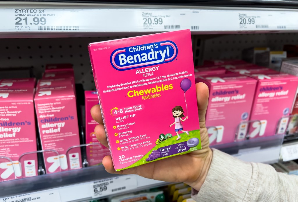 A shopper's hand holds a box of Children's Benadryl grape-flavored chewable antihistamine tablets in a pharmacy aisle.