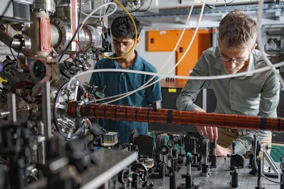 Two men stand behind a tabletop full of mirrors, lenses and lasers. 
