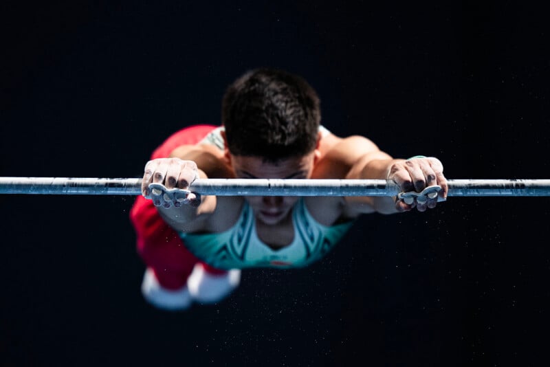 A gymnast grips a horizontal bar with chalked hands, mid-routine, wearing a teal and red uniform against a dark background.