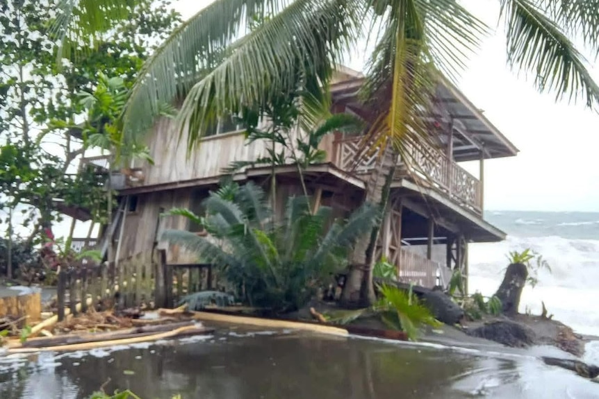 A two-storey house sits lopsided on the foreshore as waves lap at its front