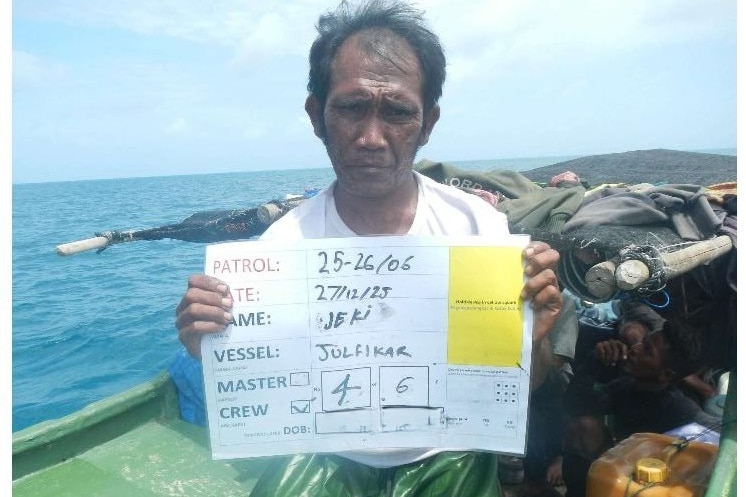 A man in a white shirt sits facing the camera holding a sign with his personal details written on it.