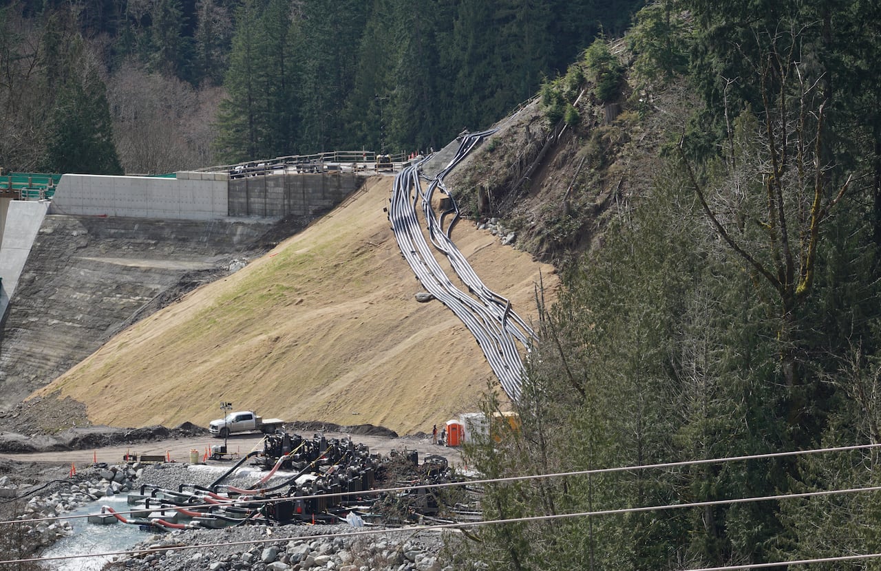 A system of pipes run overtop of a grassy hill, beside a giant concrete dam.
