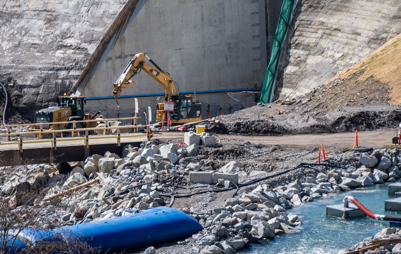 Construction vehicles work at the base of a massive concrete dam.