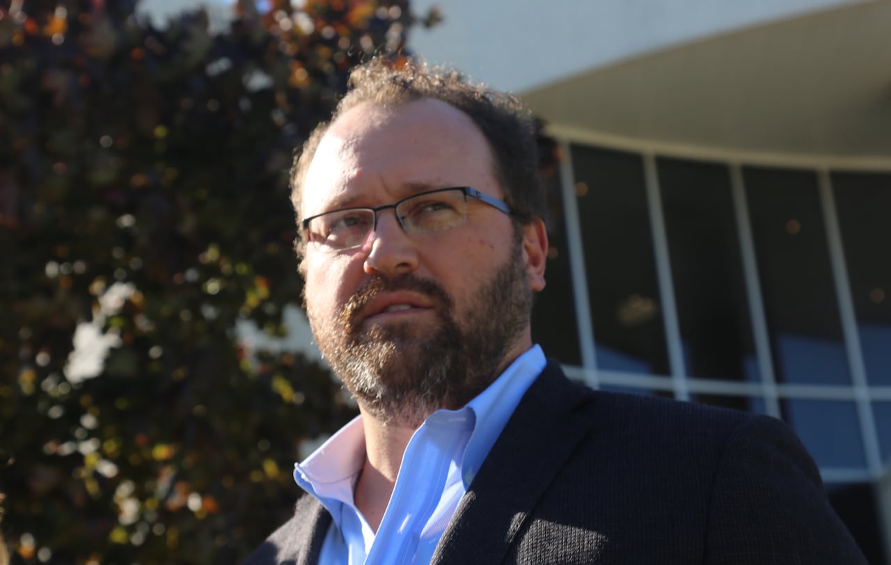 A white man with glasses and a beard stands outside a building.