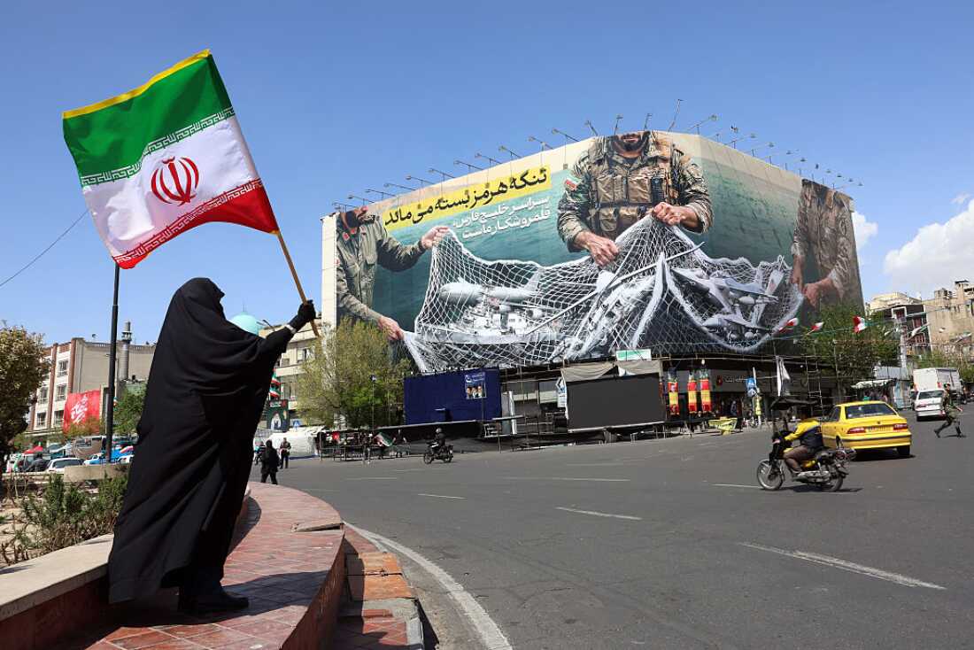 TOPSHOT - A woman holds Iran's national flag while standing near a billboard with a sentence reading 'The Strait of Hormuz remains closed' at the Enqelab Square in Tehran, on April 5, 2026.
