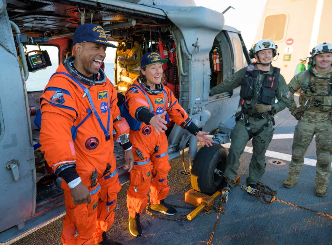 NASA astronaut Victor Glover, Artemis II pilot, left, and NASA astronaut Christina Koch, Artemis II mission specialist are seen sitting on a Navy MH-60 Seahawk from Helicopter Sea Combat Squadron (HSC) 23 on the flight deck of USS John P. Murtha.