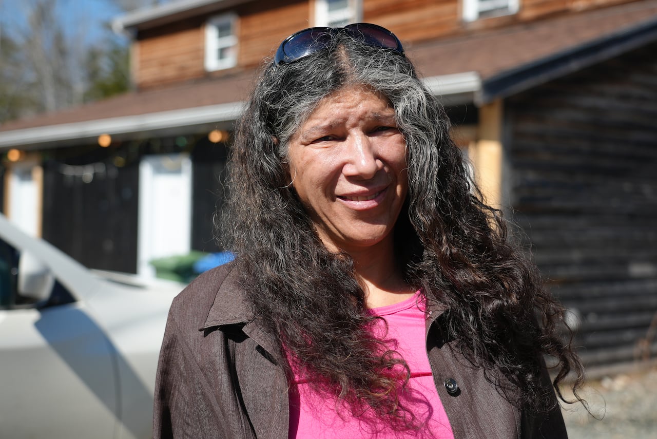 woman in pink shirt with brown jacket stands near a house