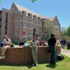 In this photo, Florida State University students wait outdoors, some checking their phones, amid an active shooter incident. A multistory brick building is in the background.