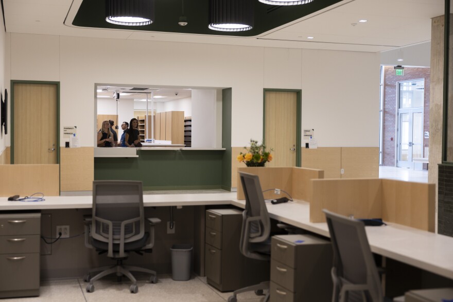 A desk area and chairs sit empty in a new health building. Beyond them, a pharmacy counter is visible.