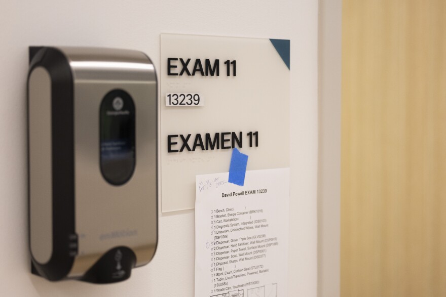 A handspray device sits on the wall outside of an exam room. A paper with information about the exam room's inventory is taped to the Exam Room sign.