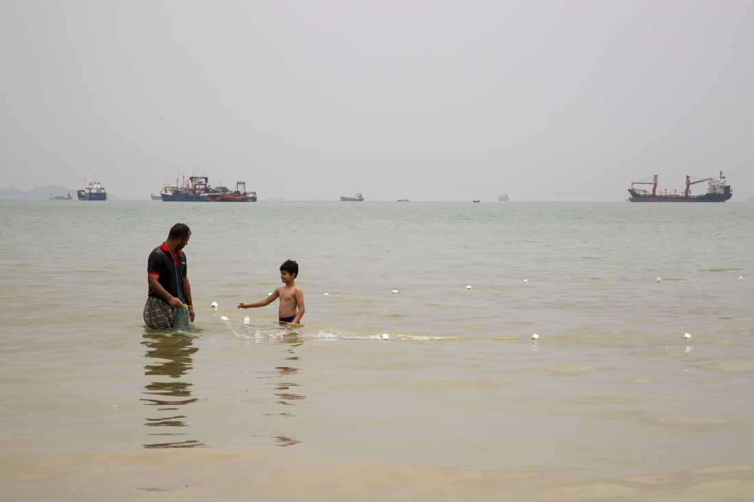 A boy walks near a man with a fishing net as ships are anchored near the shoreline in Bandar Abbas, an Iranian port city and the capital of Hormozgan province, along the Persian Gulf and Strait of Hormuz.