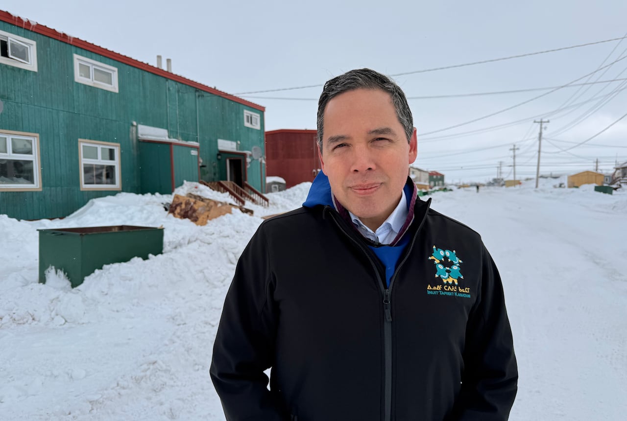 A man with dark hair, wearing a black jacket adorned with a crest, stands on a snow-covered street.