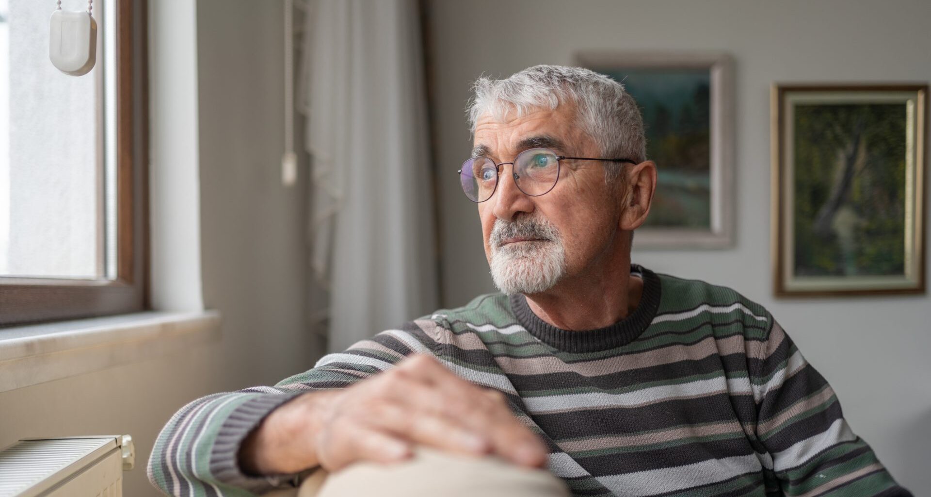 Senior Man Sitting On Sofa By Window And Looking Outside Through Window At Home.