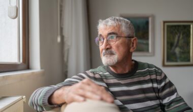 Senior Man Sitting On Sofa By Window And Looking Outside Through Window At Home.
