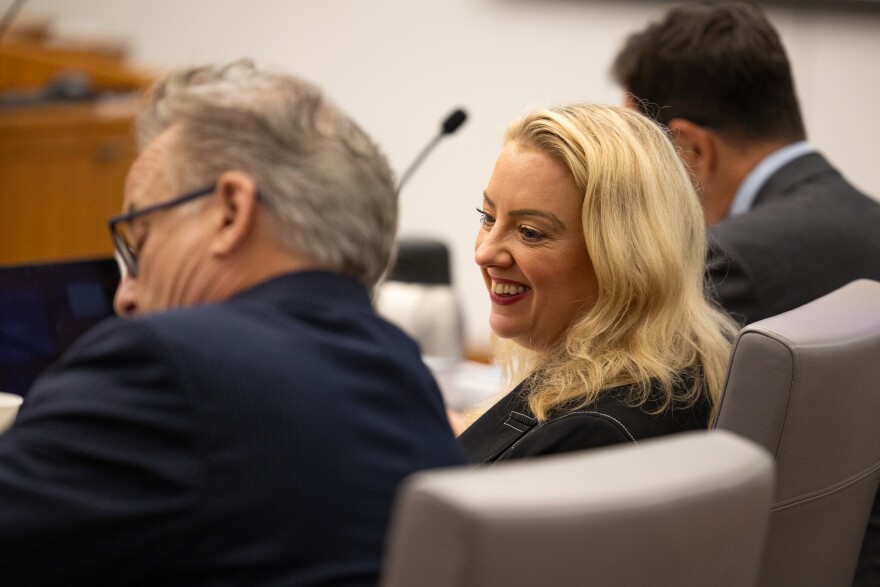 A woman seated in a chair near the front of the courtroom smiles at a colleague seated beside her.