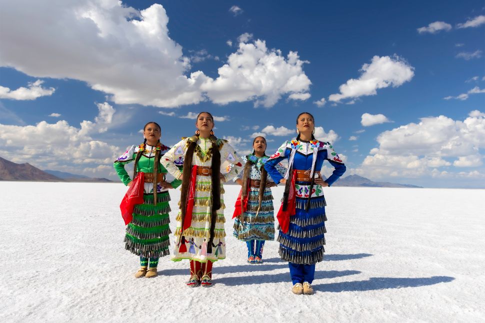 A group of four young Native American women in colorful traditional dress stand on a white salt flat landscape under a bright blue sky with scattered clouds.