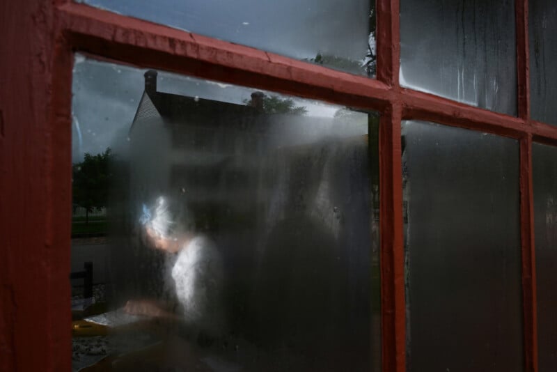 A person is seen through a foggy windowpane with red frames, their figure blurred by condensation. A house and some trees are visible outside, faintly reflected in the glass.