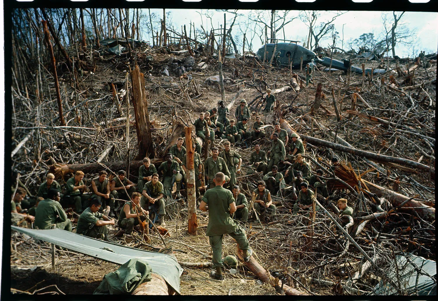 A group of about three dozen soldiers in green fatigues rest on a hillside stripped of most of its vegetation. Shattered tree trunks and fallen branches litter the ground, and a military helicopter is visible in the background.