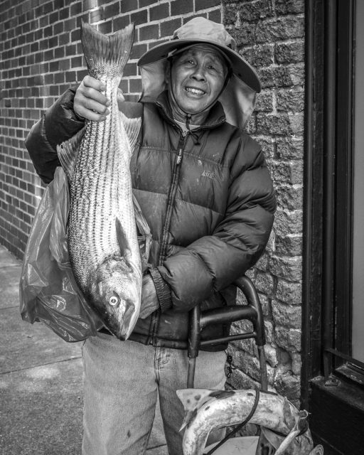 An older man in a hat and puffy jacket holds a large striped fish in one hand and stands next to a walker on a sidewalk.