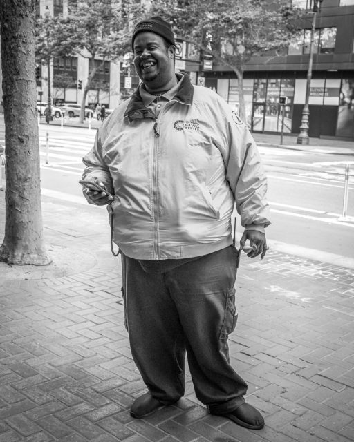 A man wearing a City Street Guides jacket stands on a sidewalk, holding a lanyard and smiling, with buildings and trees in the background.