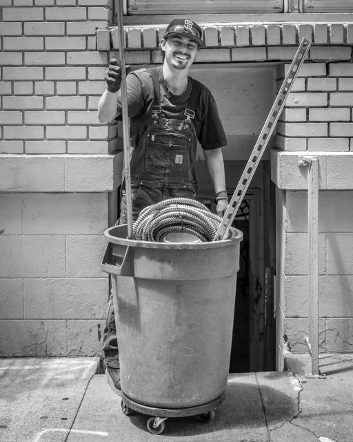 A man wearing overalls and a cap stands behind a large garbage bin on wheels, holding a long pole and smiling at the camera. Coiled tubing is visible in the bin.