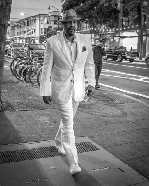 A man in a white suit, white shoes, and a hat walks down a city sidewalk at dusk; bicycles and cars are visible in the background.