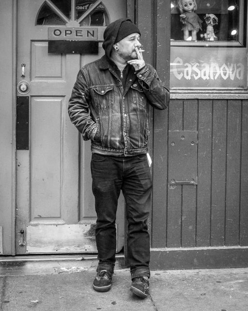 A man in a denim jacket and beanie smokes a cigarette while standing outside a door labeled "open" next to a window with "Casanova" written on it.