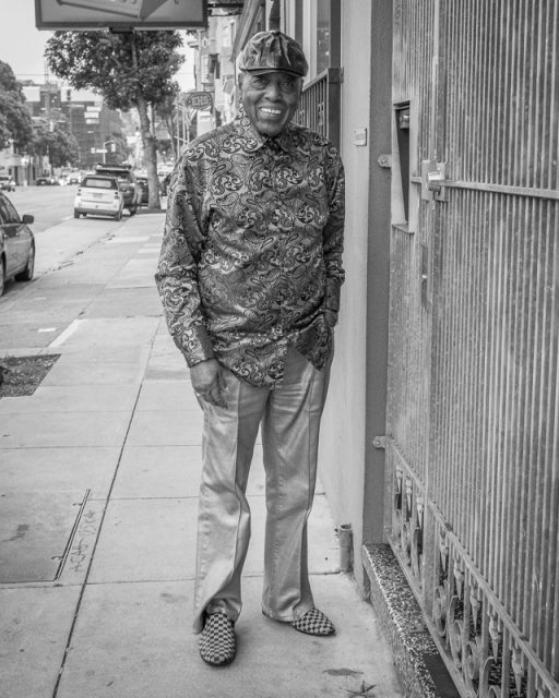 A man wearing a patterned shirt, matching cap, and patterned shoes stands on a city sidewalk next to a building, smiling at the camera. The photo is in black and white.