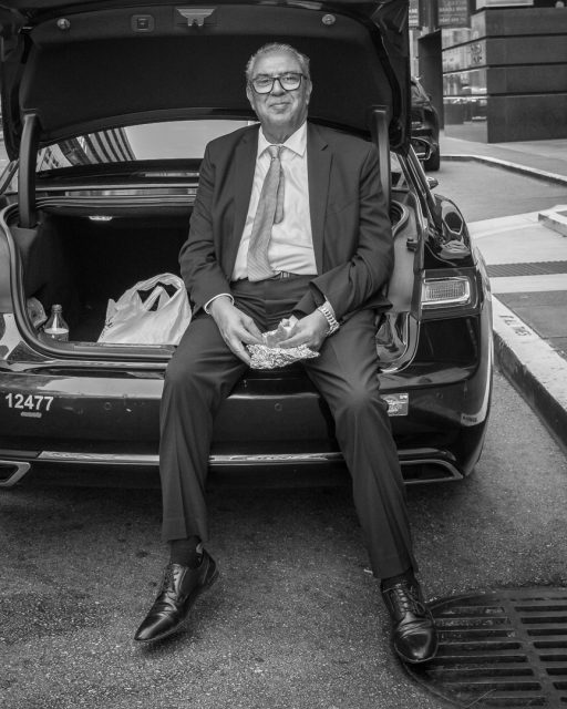 A man in a suit sits on the open trunk of a car, holding food in his hands, with shopping bags and items visible in the trunk on a city street.
