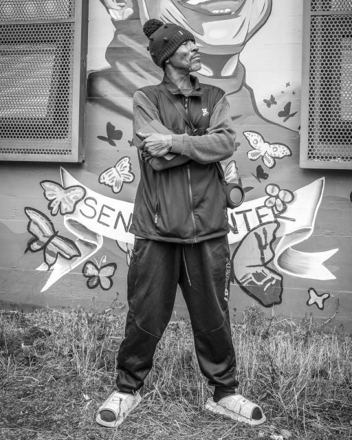 A man in casual clothing and sandals stands with arms crossed in front of a mural featuring butterflies and the word "SEN" visible. The image is in black and white.