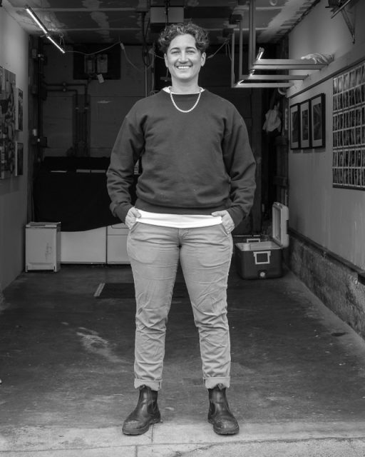 A person with short curly hair stands smiling in a garage, wearing a dark sweater, light pants, and boots, with hands in pockets. Black and white photo.