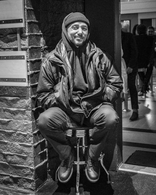 A man wearing a beanie, hoodie, and jacket sits on a stool by a brick wall indoors, looking at the camera and smiling slightly. The image is in black and white.