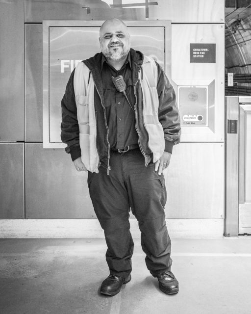 A security guard stands indoors, wearing a uniform, reflective vest, walkie-talkie, and boots, in front of a wall with signs and emergency equipment.