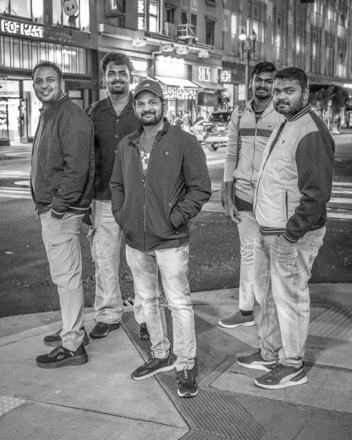 Five men stand on a city street corner at night, facing the camera and smiling; illuminated store signs and buildings are visible in the background.