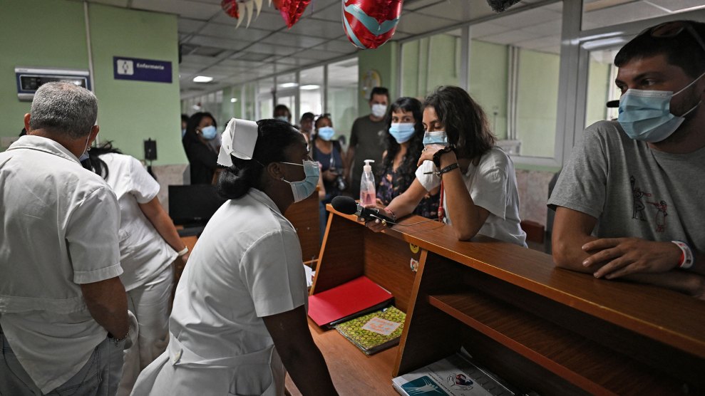A person in a white medical uniform and nurse's cap stands behind a wooden counter, speaking into a microphone held by another person. Several people wearing blue surgical masks stand on both sides of the counter and in the background of a hallway. A sign on the wall behind the counter reads "Enfermería" with a medical symbol. Red and white heart-shaped balloons hang from the ceiling. A person on the right, wearing a grey t-shirt and a blue surgical mask, leans against the counter looking toward the person in the medical uniform.