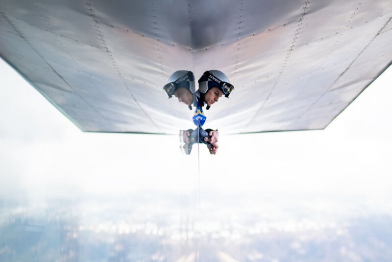 A person wearing a helmet and goggles leans out from under the metallic wing of an aircraft, holding a camera, with their reflection visible on the shiny surface above. The background is a hazy sky.