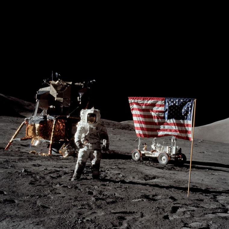 Harrison Schmitt stands on the  surface of the moon in a spacesuit, with an American flag posted in the ground on his right, and a lunar module pilot on his left