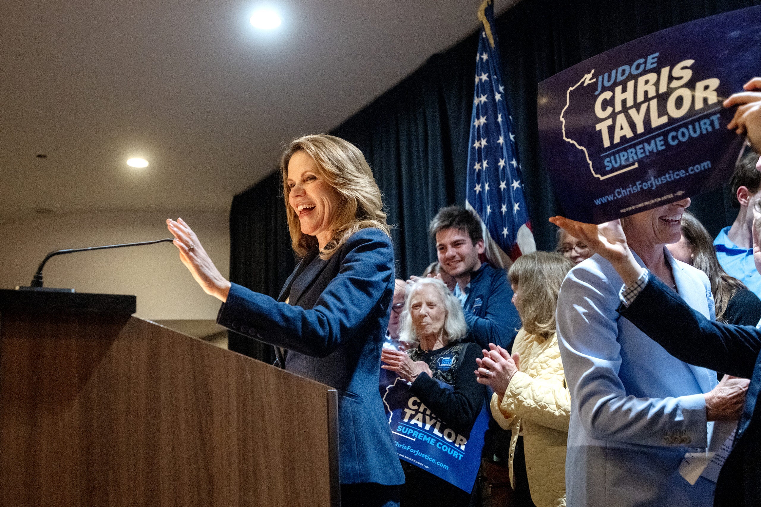 A woman speaks at a podium, smiling, as a crowd behind her holds signs reading Judge Chris Taylor Supreme Court near an American flag.
