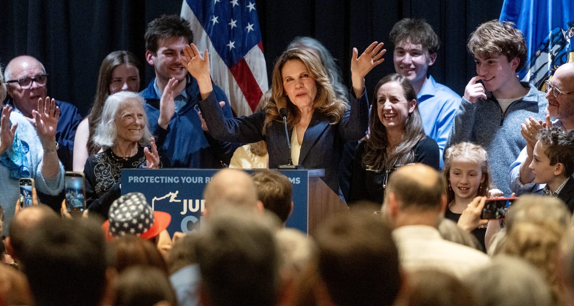 A woman at a podium addresses a cheering crowd, surrounded by supporters, with American flags in the background.
