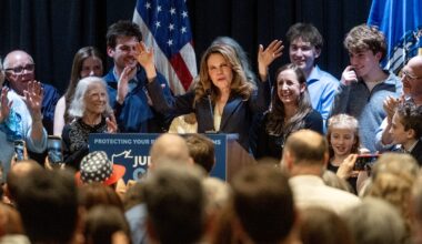 A woman at a podium addresses a cheering crowd, surrounded by supporters, with American flags in the background.